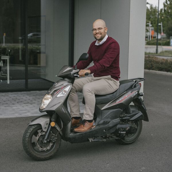 Um homem em um suéter bordô e calças bege está sorrindo em um scooter preto, estacionado em frente a um edifício de escritórios.