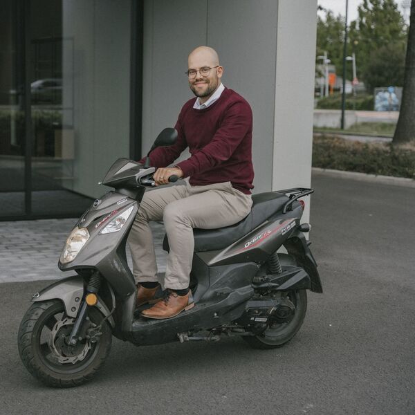 Een man in een bordeauxrode trui en beige broek zit glimlachend op een zwarte scooter, geparkeerd voor een kantoorgebouw.