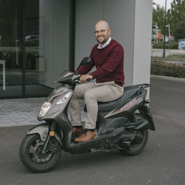 A man in a burgundy sweater and beige pants smiling while sitting on a black scooter, parked in front of an office building.