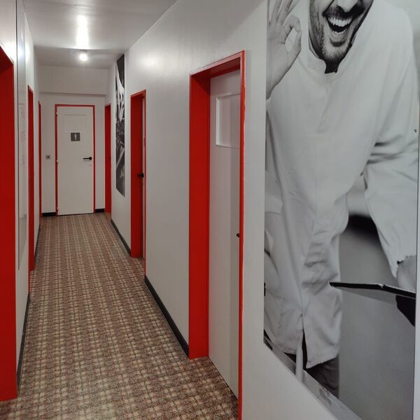 A hallway with red door frames and a large black-and-white portrait of a smiling man with a hairnet making an 'okay' hand gesture.